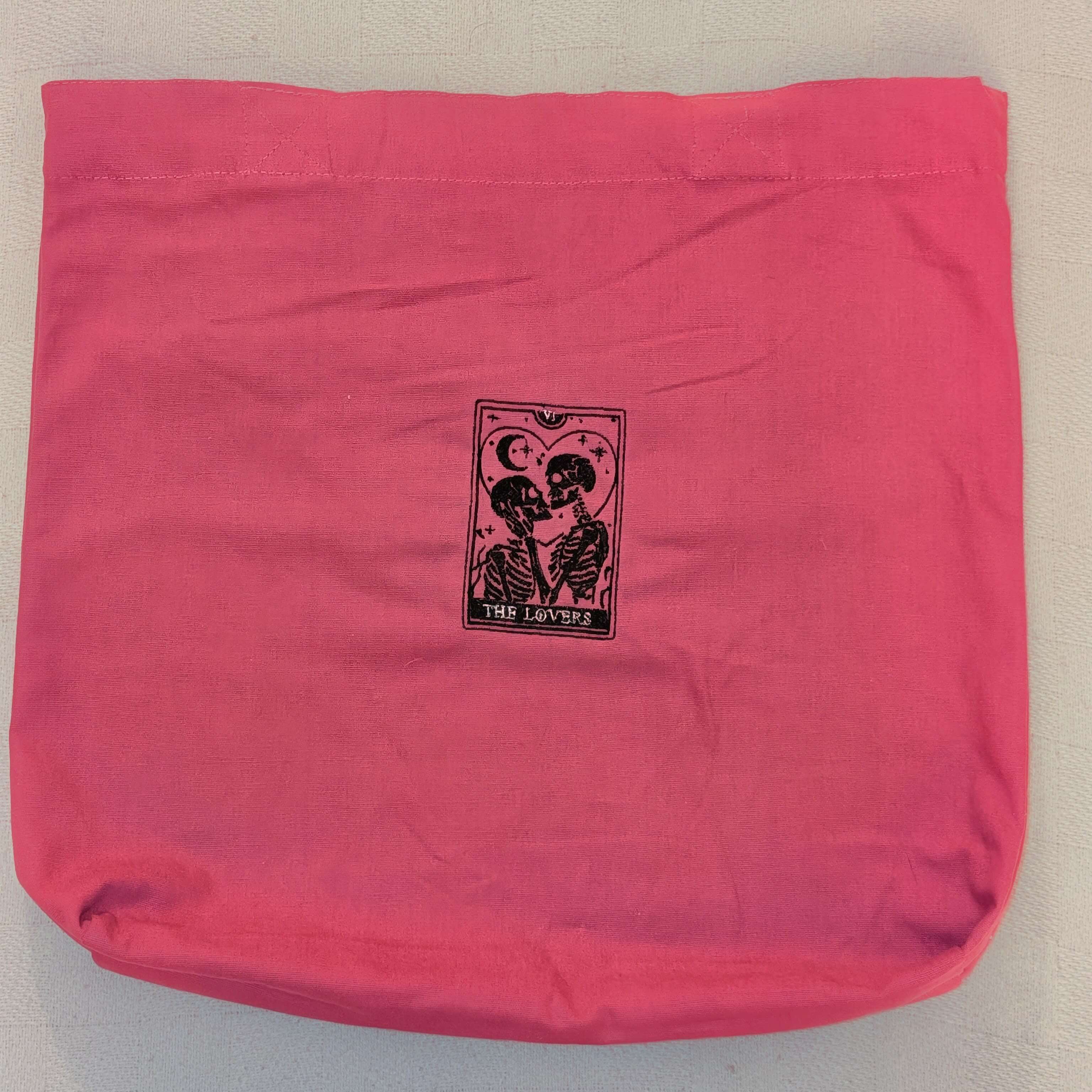 Pink canvas tote bag featuring a black tarot-style embroidery design of two skeletons embracing, titled The Lovers.